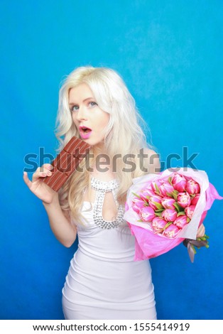 beautiful young model girl with a bouquet of pink flowers and a chocolate bar on a blue background in the Studio