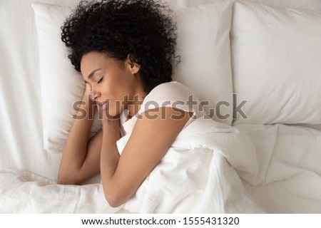 Beautiful African American woman with hands under cheek sleeping in bed under blanket top view, girl with closed eyes resting in bedroom, enjoying fresh bedclothes, lying on soft pillow close up Royalty-Free Stock Photo #1555431320