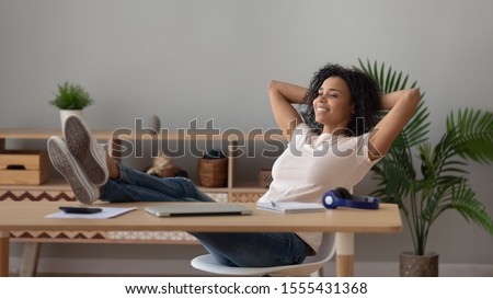 Satisfied African American woman relaxing at workplace with legs on table, happy female freelancer student dreaming, resting after work done in comfortable chair at home