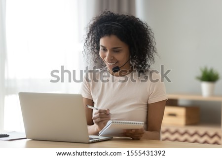 Smiling African American woman in headset writing notes in notepad, using laptop, satisfied girl watching webinar, student in headphones with microphone learning language, listening online lecture
