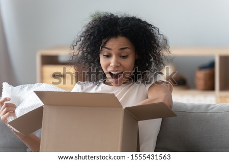 Surprised African American woman unpacking parcel at home close up, amazed girl with wide open eyes and mouth looking in cardboard box, satisfied client received online store order, delivery concept Royalty-Free Stock Photo #1555431563