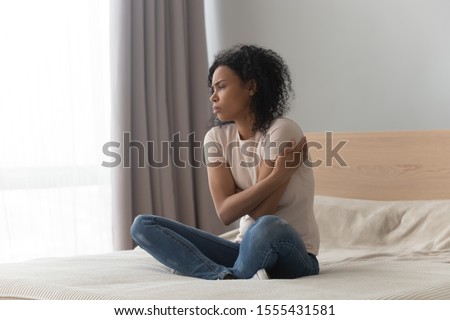 Upset sad African American woman sitting in bed alone, hugging herself, feeling lonely, unhappy girl has psychological troubles, trauma, thinking about problem in bedroom, unwanted pregnancy Royalty-Free Stock Photo #1555431581