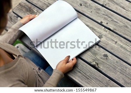 Young woman sitting at the table with a booklet with white pages. The white pages can be used for any logos, label signs or any graphic additions.