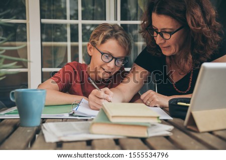 Young student doing homework at home with school books, newspaper and digital pad helped by his mother. Mum writing on the copybook teaching his son. Education, family lifestyle, homeschooling concept Royalty-Free Stock Photo #1555534796
