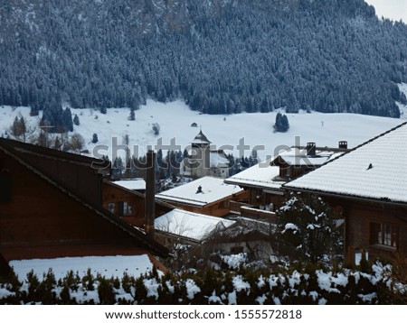 A small simple church in a snowy alpine village full of chalets with a pine forest in the background.