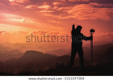 Silhouette portrait  image of photographer man holding camera with tripod with warm tone morning light and point to the sky over mountain complexity layer background .