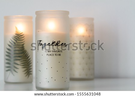Three lit candles with design on their glass, one in focus and in front of others, on a white table and background with copy space