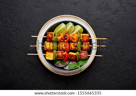 Paneer Tikka at skewers in black bowl at dark slate background. Paneer tikka is an indian cuisine dish with grilled paneer cheese with vegetables and spices. Indian food. Top view Royalty-Free Stock Photo #1555665335