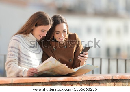 Two happy tourists checking paper map and mobile phone in a balcony on winter vacation