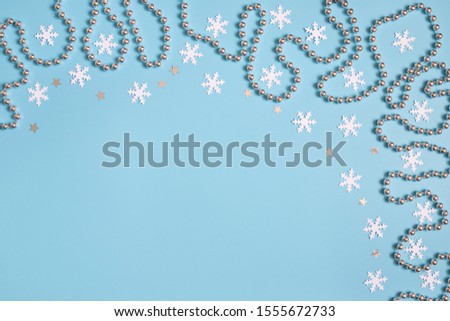 Christmas border with silver garland and stars, white snowflakes on pastel blue background. Space for text. Top view, flat lay. Copy space. Greeting card. Winter concept.