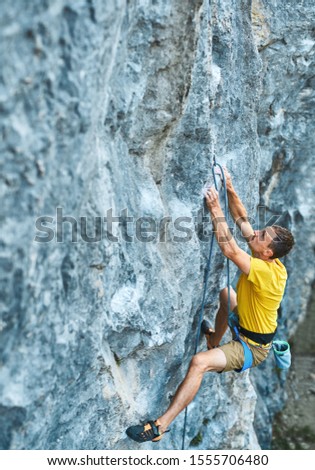 young strong man rock climber climbing on a high vertical limestone cliff, reaching holds, making hard wide move and gripping hold, attaching rope. Conquering, overcoming and active lifestyle concept.