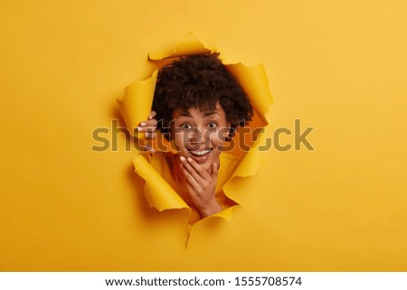 Merry brunette adult woman holds chin, smiles broadly, shows good dental treatment, has healthy skin, looks curiously happily through ripped paper hole, yellow bright background, has fun alone