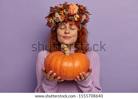Horizontal shot of pretty redhead woman closes eyes with pleasure, holds ripe orange pumpkin, demonstrates autumn harvest, wears creative handmade wreath on head. Season, people, vegetables.