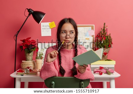 Serious contemplating Asian girl with combed plaits, holds notebook and pencil, writes down future plans and goals for coming New Year holidays, poses with own diary in study room over rosy wall Royalty-Free Stock Photo #1555710917