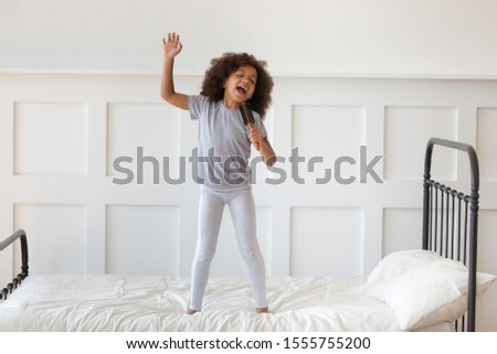 Cute little preschooler african American girl stand on bed sing in toy hairbrush microphone, funny small artistic biracial kid have fun playing at home in morning, perform entertain in bedroom