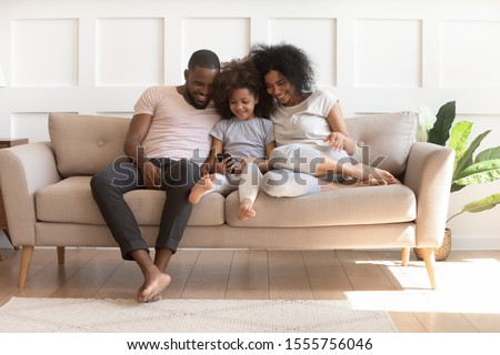 Happy young african american family sit on couch watch cartoons on smartphone at home together, smiling loving biracial parents enjoy time play on cellphone with cute little preschooler daughter