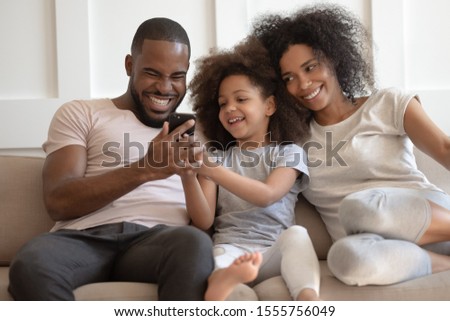 Smiling african American family sit on couch at home laugh watch funny video on cellphone, cute little girl child show smartphone to happy dad, loving mixed race parents enjoy weekend with daughter Royalty-Free Stock Photo #1555756049