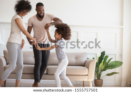 Happy african American young parents dance with cute little preschooler daughter in living room, smiling loving family with small kid have fun playing together at home, enagaged in physical activity Royalty-Free Stock Photo #1555757501