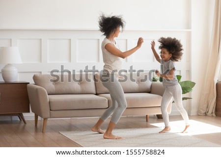 Excited young african American mom or nanny play dancing with funny little preschooler girl child, happy biracial mother parent have fun entertain with small daughter jump move to rhythm at home