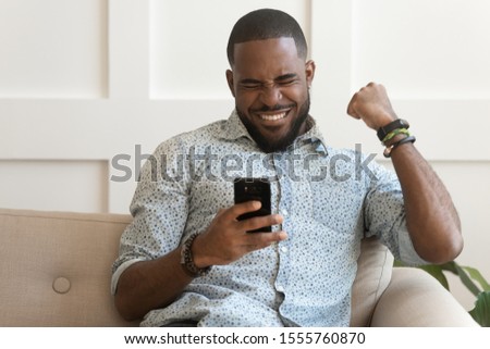 Excited african American millennial male feel euphoric winning online lottery or game on cellphone, overjoyed biracial man triumph get good pleasant text message on smartphone, reward concept Royalty-Free Stock Photo #1555760870