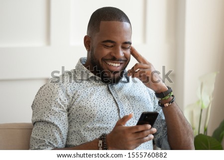 Smiling african American millennial man sit on couch at home texting messaging on smartphone, happy biracial young male relax use cellphone, browse internet, chat having pleasant conversation Royalty-Free Stock Photo #1555760882