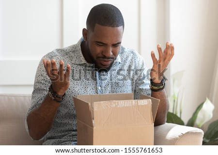 Frustrated african American millennial male buyer unpack unbox cardboard box delivery package disappointed with product quality, confused biracial man shop online open box get wrong order Royalty-Free Stock Photo #1555761653