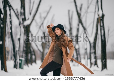 Young cheerful girl in brown stylish coat posing in winter tree valley.