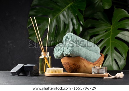 Spa and bath concept. Green tea scrub, coal black soap, eucalyptus, oil, sea stones, towels and body brush on black background with tropical leaves. Copy space. Royalty-Free Stock Photo #1555796729