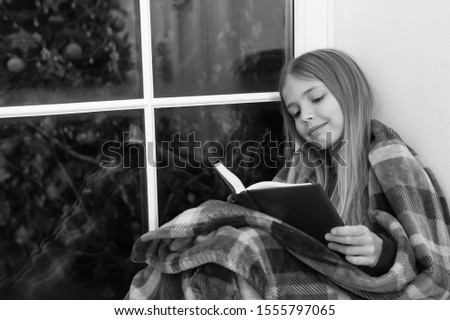 Fairy tale for merry holidays. Little reader wrapped in plaid sit on window sill. Little girl enjoy reading Christmas story. Little child read book on Christmas eve. Childrens book. Magic xmas spirit.