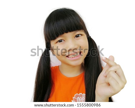 portrait of smiling asian child ​girl​ wear orange shirt​ show mini heart sign with her hand, standing on​ over white​ background, happiness time concept, selective focus on face 