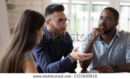 In board room gathered multi racial business people focus on serious middle eastern ethnicity team leader talking about strategy, corporate goals, share ideas, solve problems together at group meeting Royalty-Free Stock Photo #1555830818