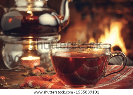 Tea with hawthorn in a glass cup and teapot with a tealight on a wooden table in a room with a burning fireplace