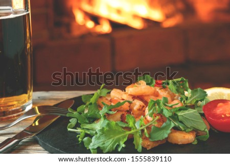 Fresh homemade salad of shrimp, arugula and tomato on a black slate dish and beer on a wooden table in a room with a fireplace, close-up