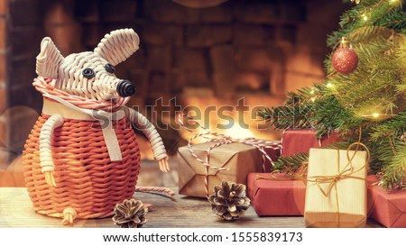 Christmas composition - a wicker white mouse - symbol of 2020 according to Chinese horoscope next to gifts under Christmas tree in room by fireplace.
