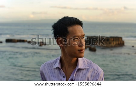 An Asian Thai man tilts his head to one side looking contemplative thoughtful and melancholic as if he is pondering about the future, with the sea of Bali Indonesia as the background Royalty-Free Stock Photo #1555896644