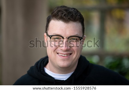 A young caucasian male wearing glasses, a white t-shirt and a black hoodie smiles and looks happy and approachable.