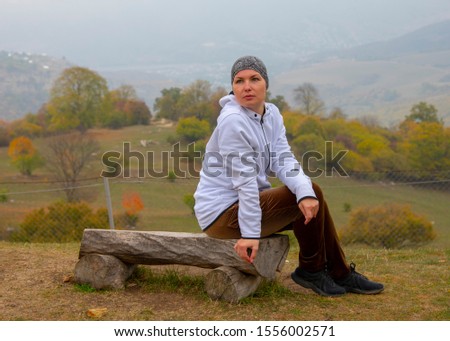 A woman in a bandana and a white hoodie sits on a wooden bench against a misty autumn landscape.