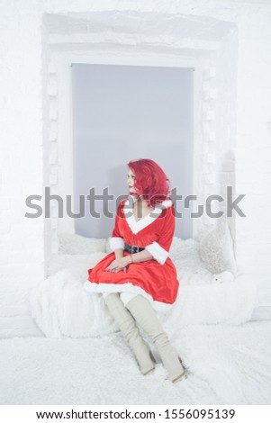 pretty redhaired young stylish woman in velvet santa dress with fur posing on white room background near the cozy window