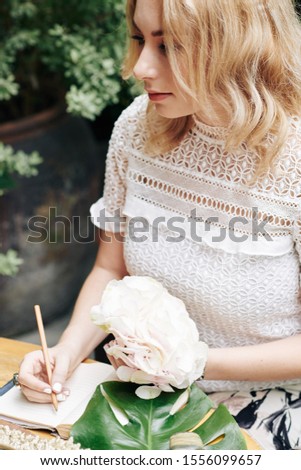Pensive young woman with beautiful flower in hand filling her gratitude journal