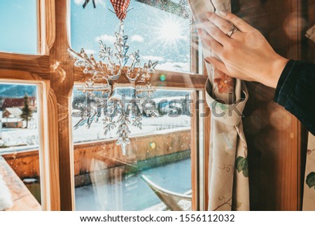 Hand Opens Curtain on the Wooden Cottage House Window. Bright Sunlight Shines Through. Beautiful snowy Winter morning light behind the Log Cabin Window. Snowflake Christmas Decoration. 