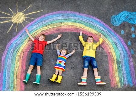 three little children, two school kids boys and toddler girl having fun with with rainbow picture drawing with colorful chalks on asphalt. Siblings in rubber boots painting on ground playing together.