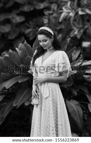 Smiling feminine lady in pink tender outfit posing at tropical plant background medium long shot. Adorable luxury young woman wearing beauty classic dress handbag and headdress looking at camera