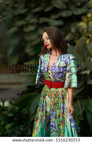 Stylish young woman wearing luxury colourful dress posing at summer garden looking at camera. Female fashion model relaxing outdoor at green tropical plant background motion body length shot