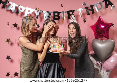 Image of a happy positive women friends isolated over pink wall background at the happy birthday party holding cake covering eyes of birthday girl.