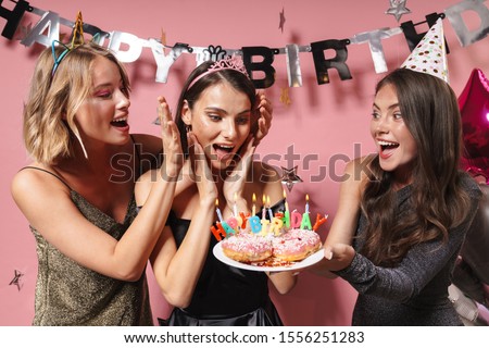 Image of a women friends isolated over pink wall background at the happy birthday party holding cake covering eyes of birthday girl.