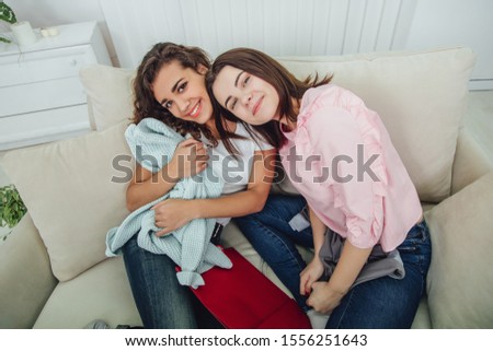 Two shopaholic girls sitting on the sofa, smiling, looking at the camera, enjoying clothes they have bought. Expressive satisfied face expressions.