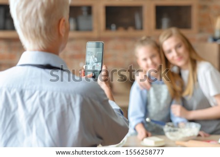 Back view of old woman taking photo of her happy daughter and granddaughter cooking in kitchen