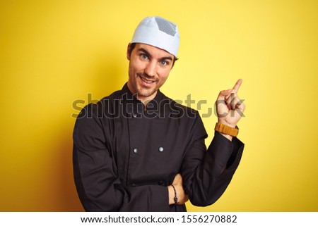 Young handsome chef man cooking wearing uniform and hat over isolated yellow background with a big smile on face, pointing with hand and finger to the side looking at the camera.