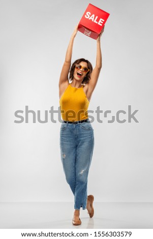 shopping and people concept - happy smiling young woman in sunglasses, mustard yellow top and jeans with sale sign posing over grey background
