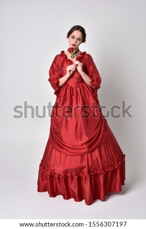 full length portrait of a brunette girl wearing a red silk victorian gown. Standing pose, holding a rose,  on a white studio background.